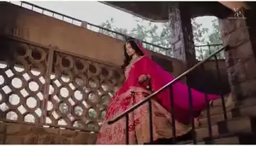 A bride in a magenta and gold lehenga with a long flowing dupatta, standing on an ornate stone balcony with a lattice screen.