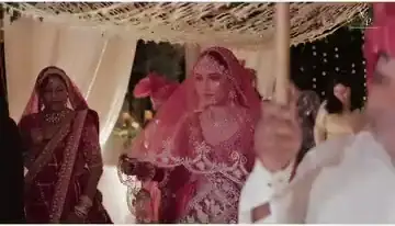A bride in a red bridal lehenga with a dupatta over her head, entering a mandap decorated with white drapes, family around her.