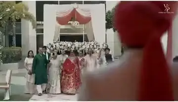 A wedding baraat: a wide shot of the couple and family walking together in front of a floral mandap, a groom's red turban in the foreground.